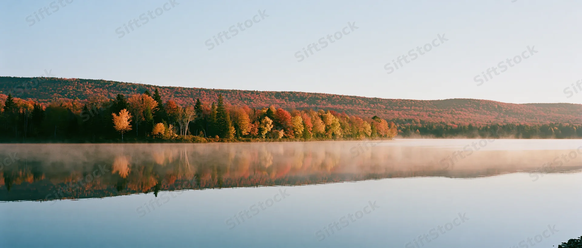 Calm lake reflecting autumn forest at dawn with morning mist over water