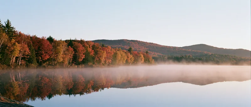 Calm lake reflecting autumn trees and mountains at dawn with morning mist
