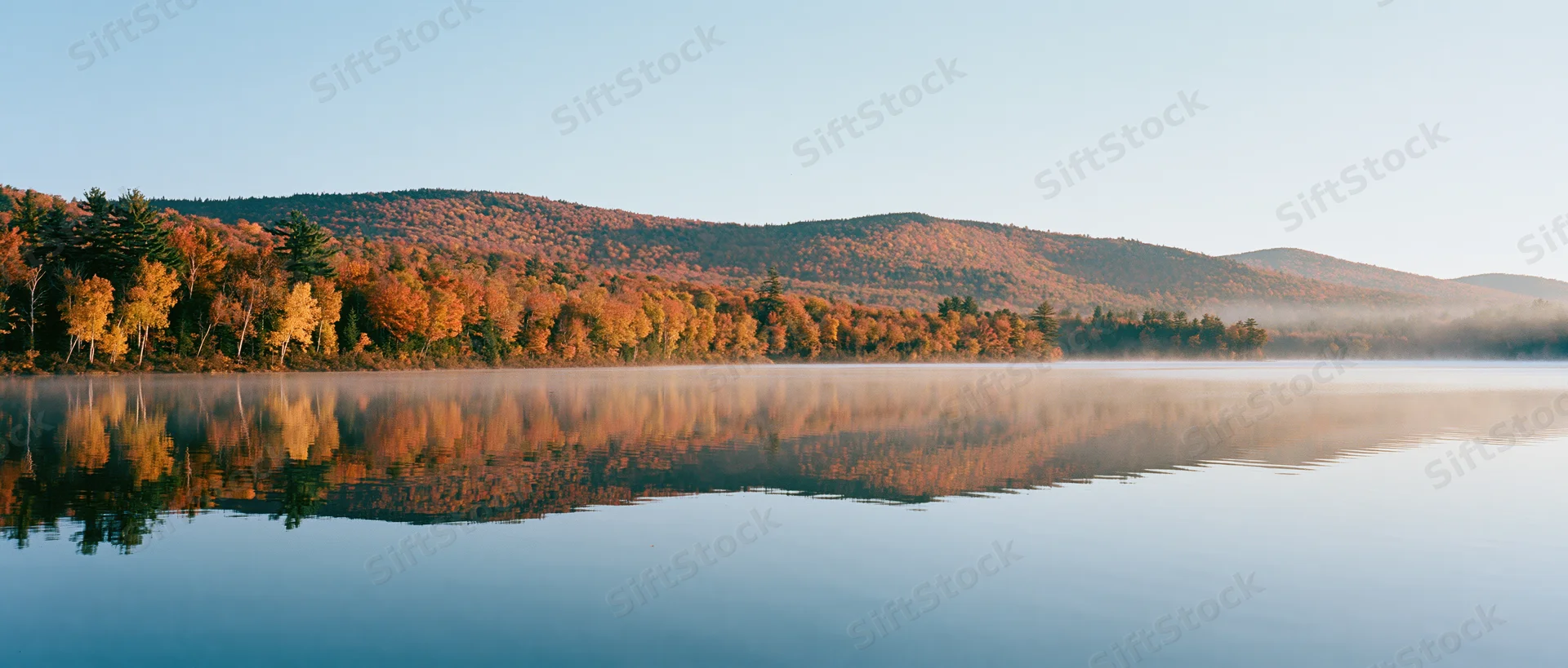 Calm lake reflecting autumn forest at dawn with mist over water and mountains in background