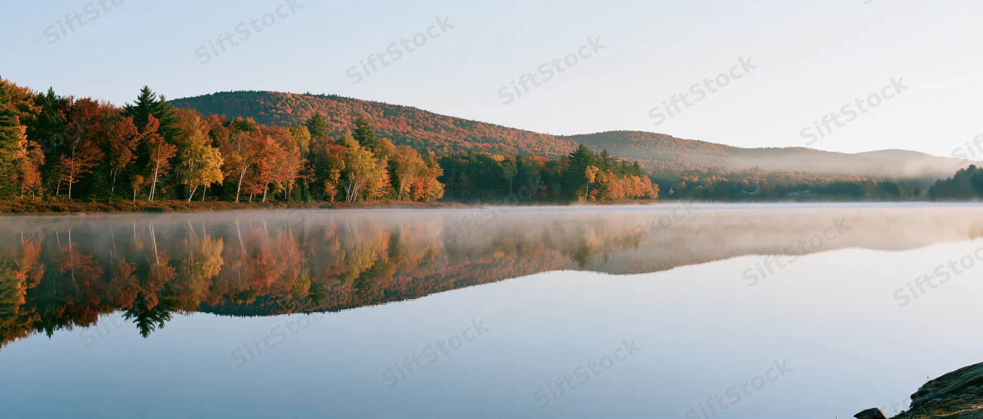 Calm lake reflecting autumn trees and hills at dawn with morning mist