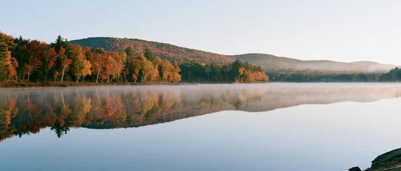 Calm lake reflecting autumn trees and hills at dawn with morning mist