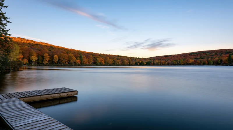 Calm lake at dusk with wooden dock extending into still water surrounded by autumn forest