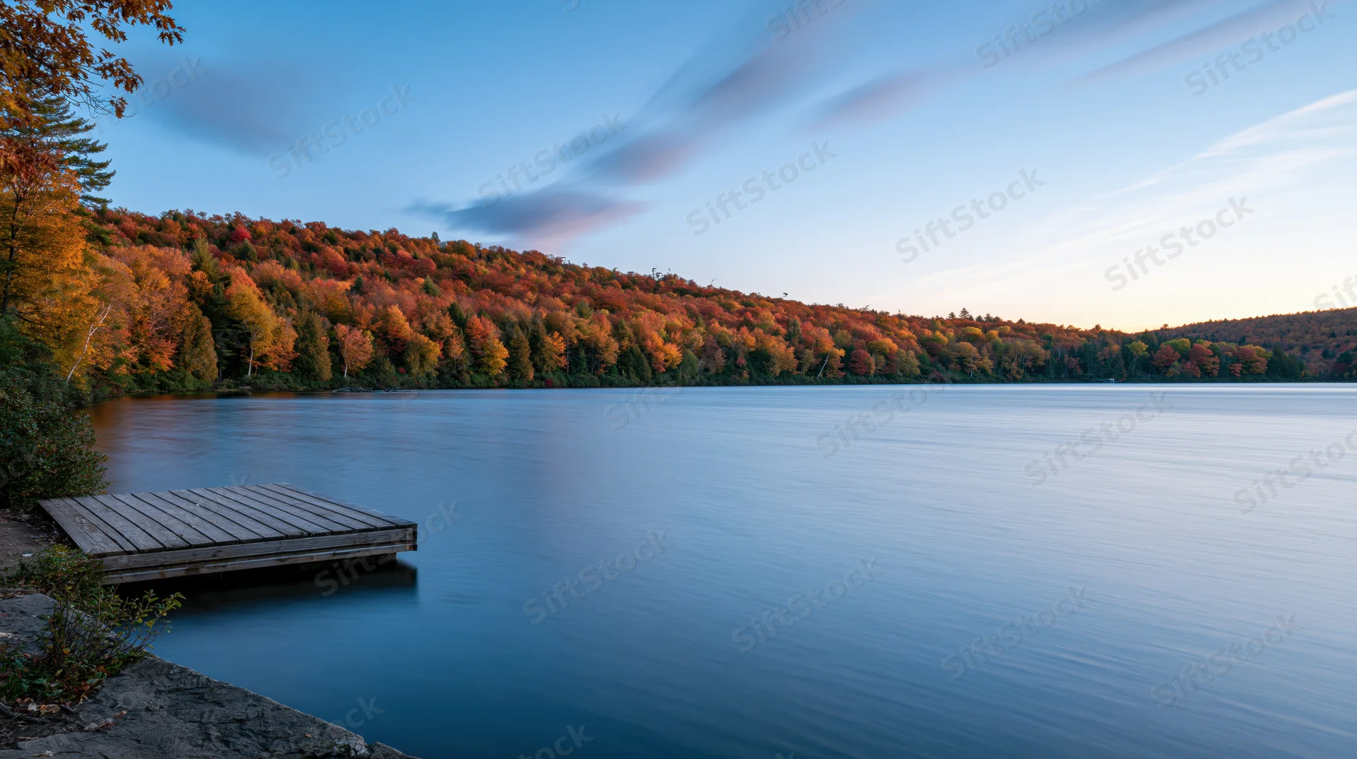 Calm lake at dusk with small wooden dock extending into water surrounded by autumn forest