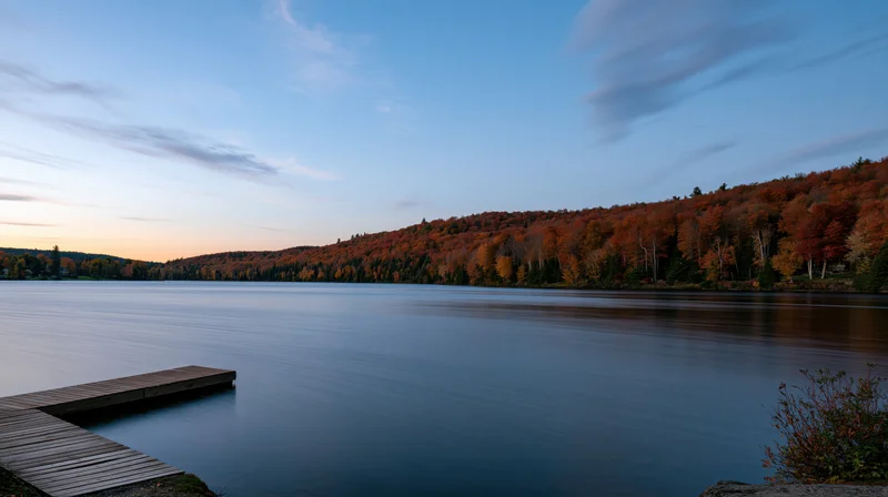 Calm lake at dusk with wooden dock extending into water surrounded by autumn forest
