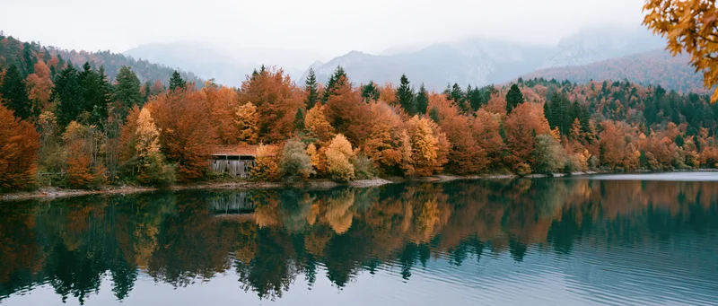 Calm mountain lake reflecting autumn trees along the shore with misty peaks in the background