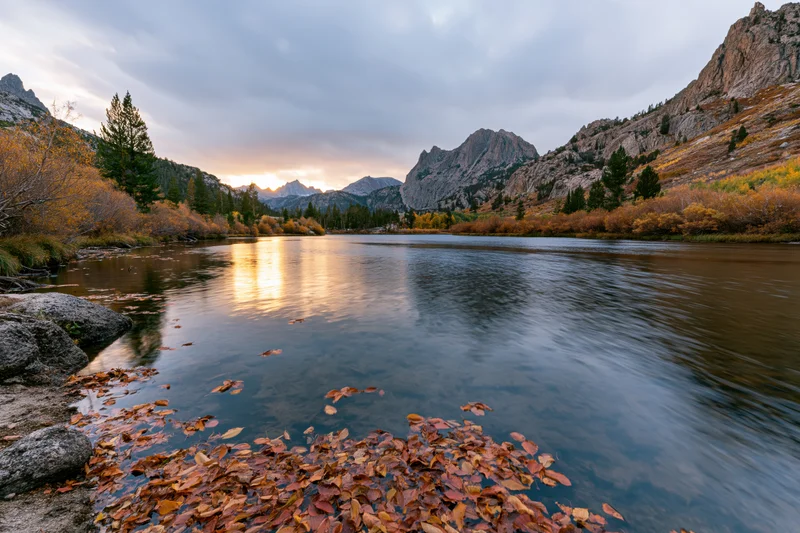 Calm river reflecting sunset light between autumn mountains with floating leaves on the shore