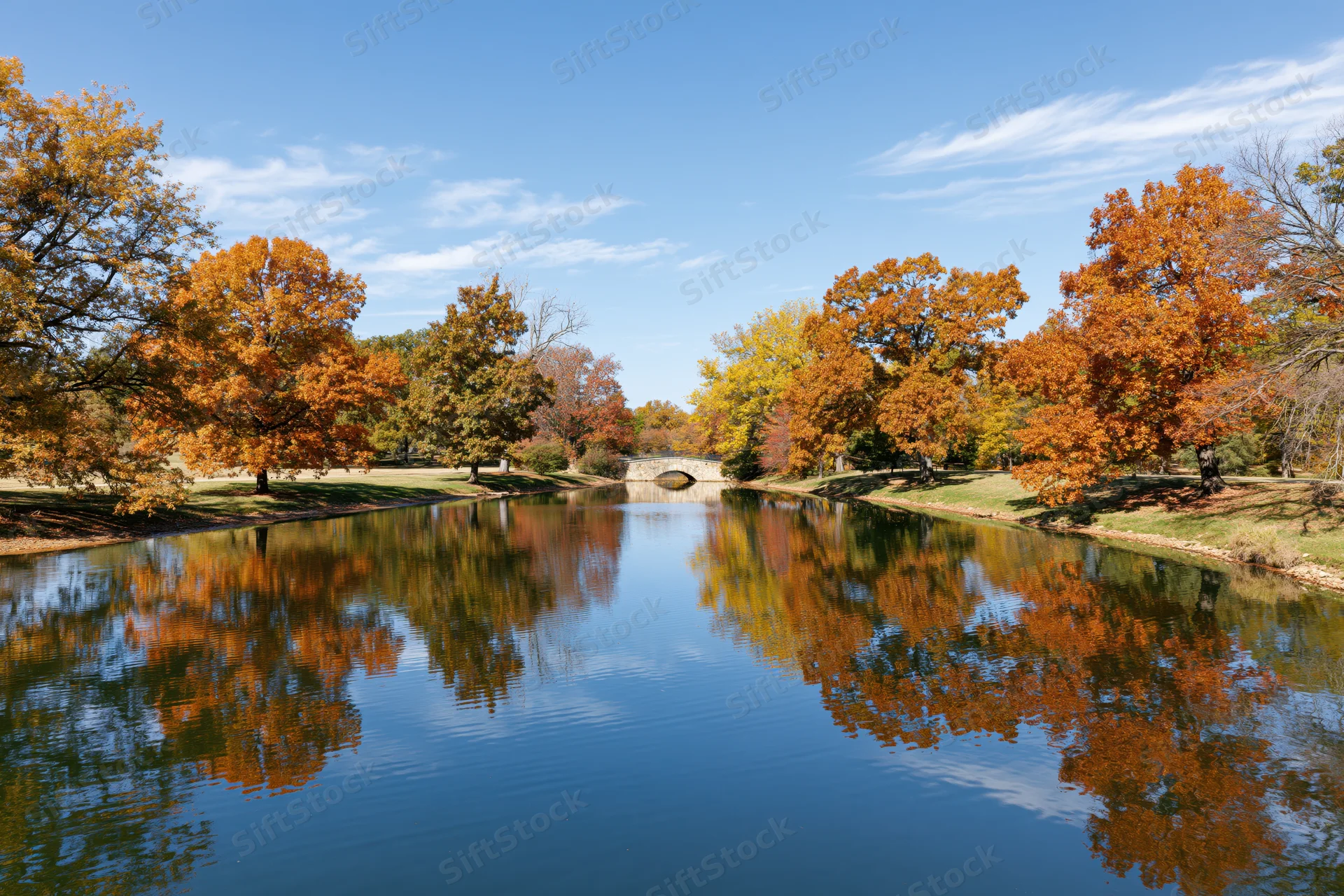 Autumn trees reflecting in calm pond at park with stone bridge in background