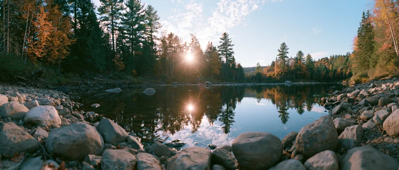 Calm shallow river reflecting autumn trees and low sun at forest edge