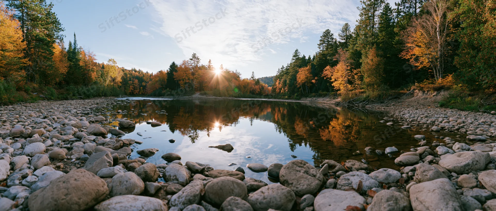 Shallow river reflecting autumn trees and low sun at sunset in forested landscape