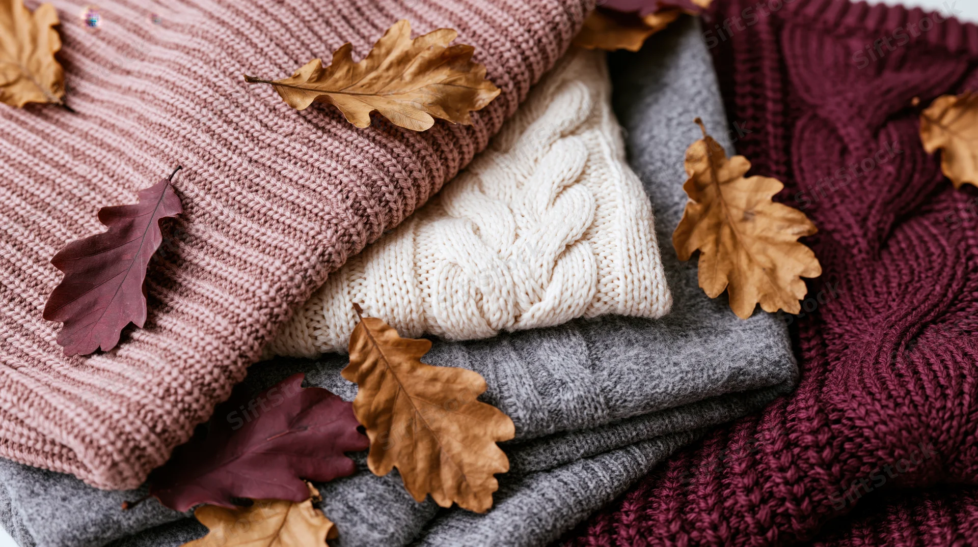 Folded knit sweaters stacked together with dried oak leaves on a flat surface