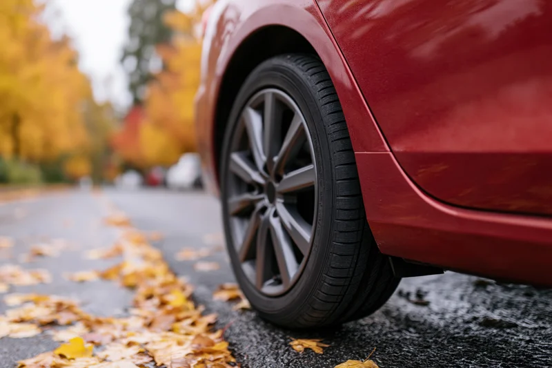 Close-up low-angle view of red sedan's front wheel and alloy rim on wet pavement with autumn leaves scattered along the road