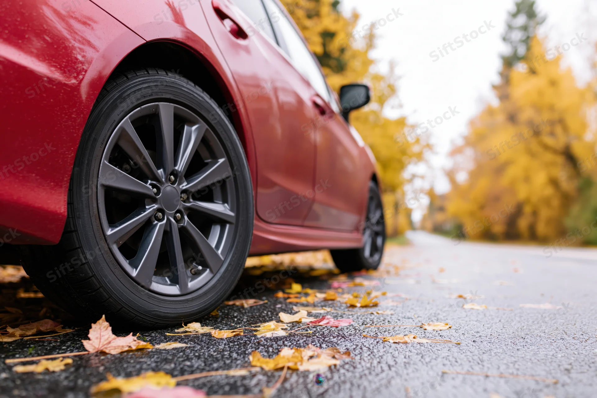 Red sedan's front wheel and alloy rim on autumn road with fallen leaves