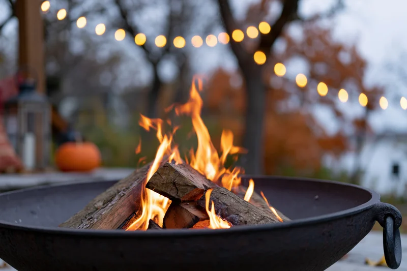 Close-up of a cast iron fire pit with burning logs and string lights in a backyard garden at evening