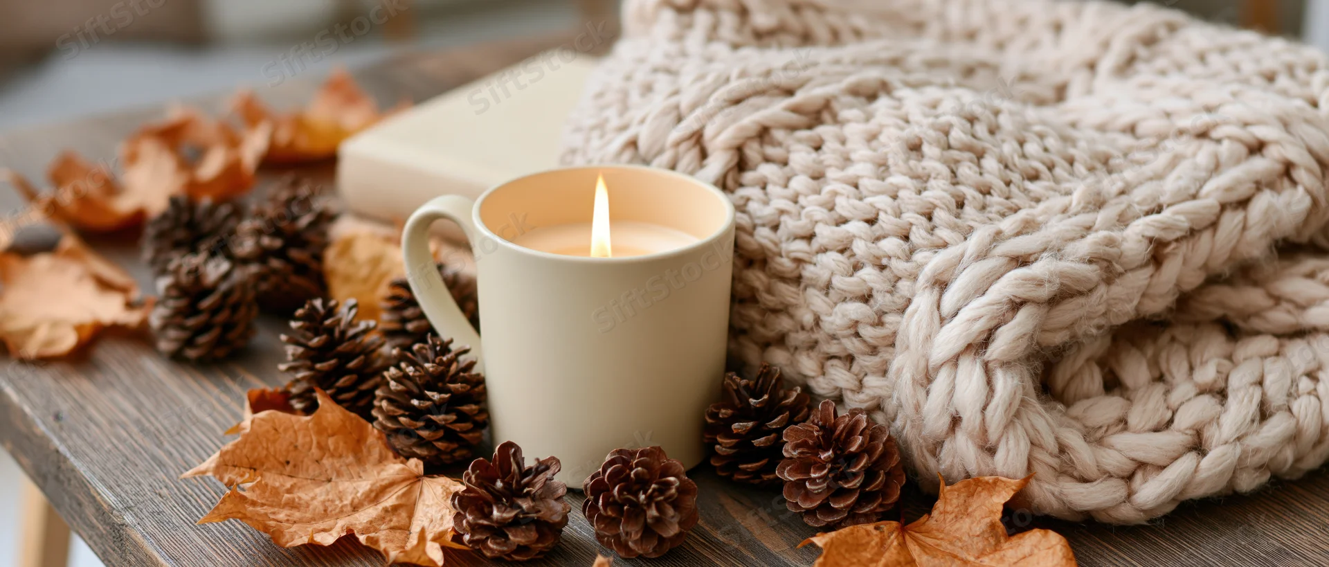Lit candle in cream mug surrounded by pinecones, dried leaves and knitted sweater on wooden table