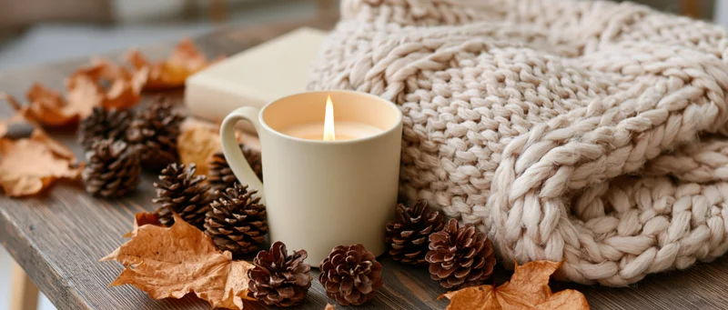 Lit candle in cream mug surrounded by pinecones, dried leaves and knitted sweater on wooden table