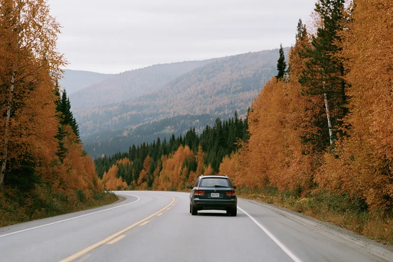 Dark suv driving away on winding two-lane highway through autumn forest with mountains in background