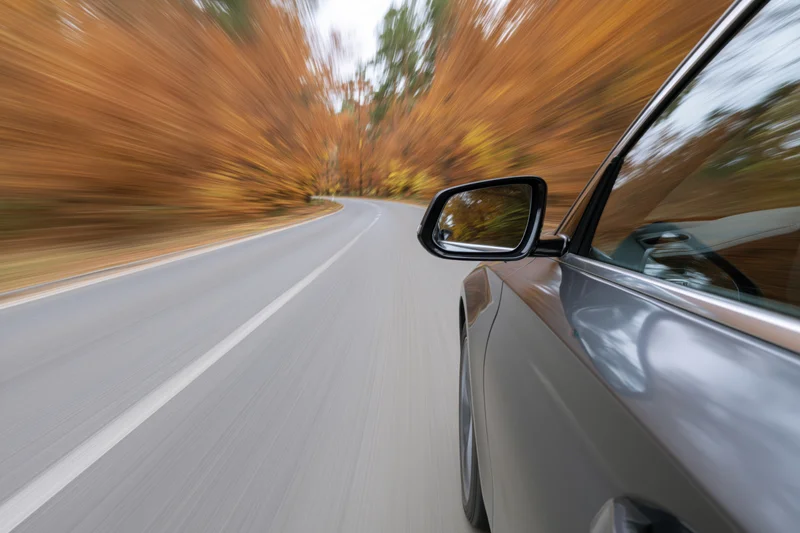 Dark silver car driving fast along a curving two-lane road lined with autumn trees