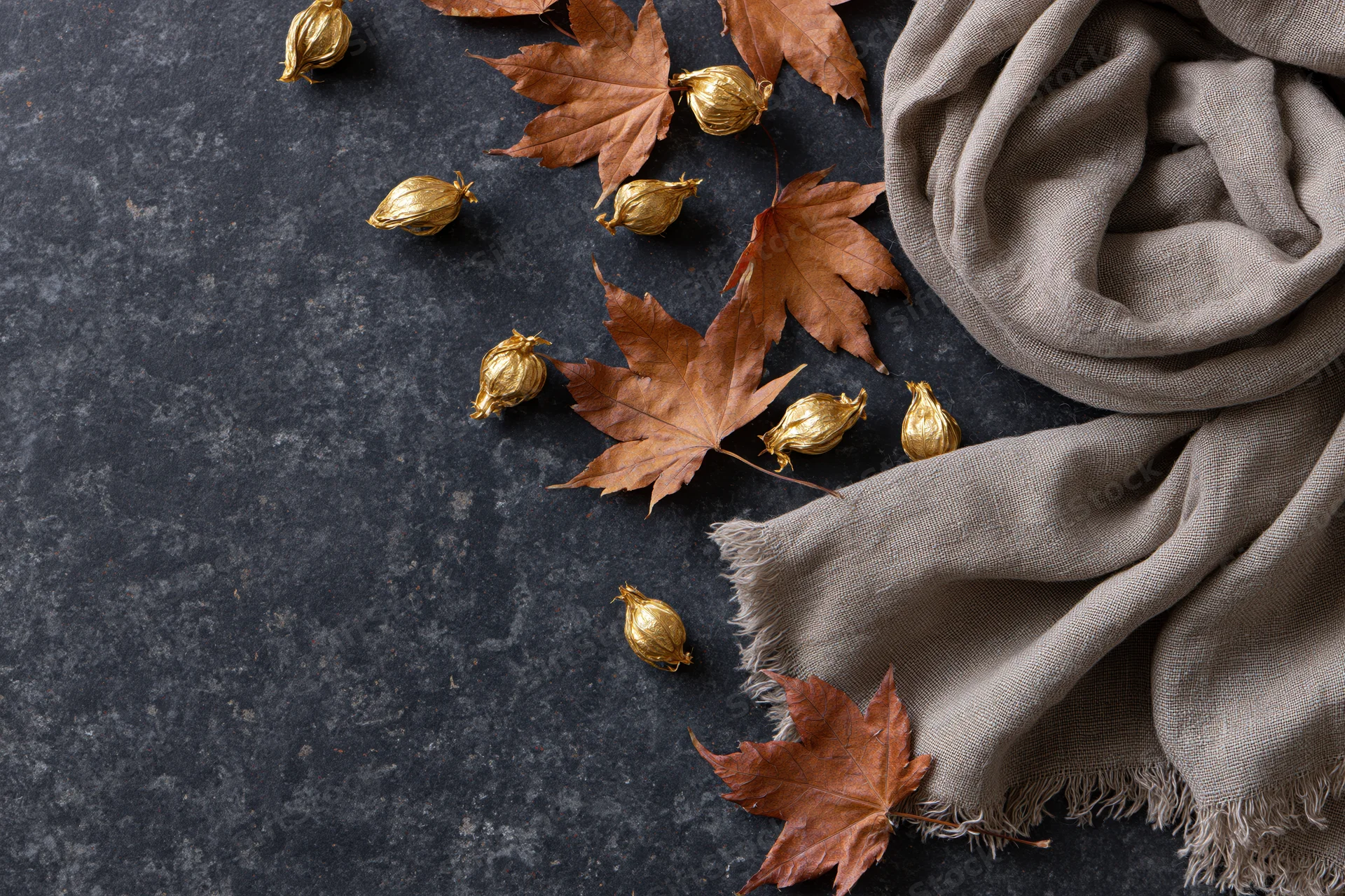 Dried maple leaves and golden seed pods arranged with a gray scarf on a dark textured surface