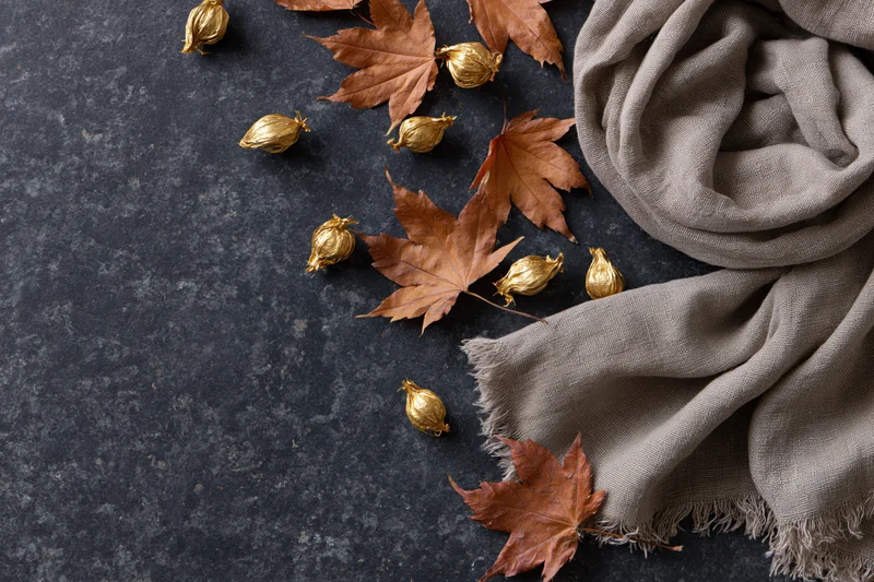 Dried maple leaves and golden seed pods arranged with a gray scarf on a dark textured surface