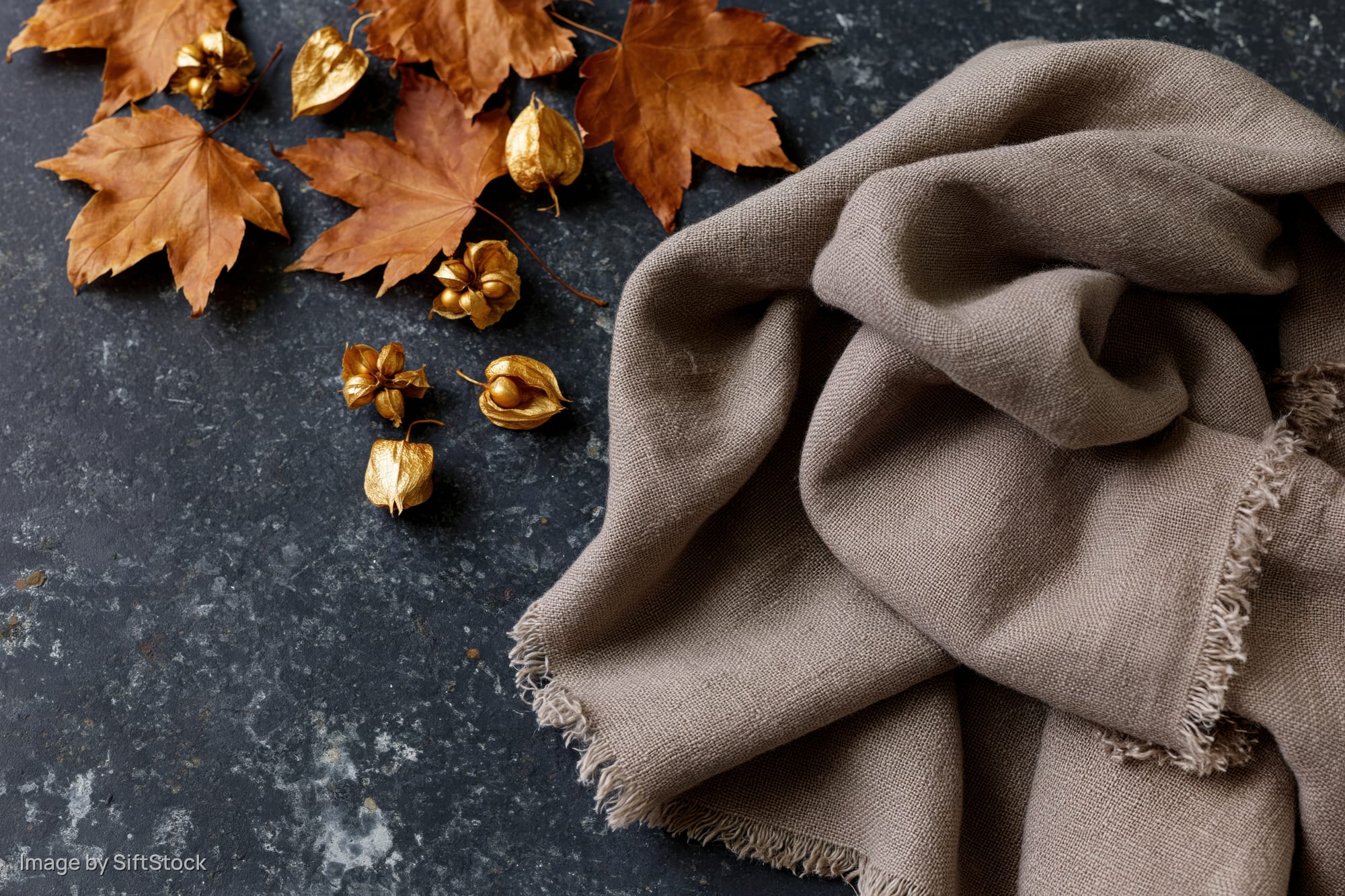 Dried maple leaves and golden seed pods arranged with a brown scarf on a dark stone surface