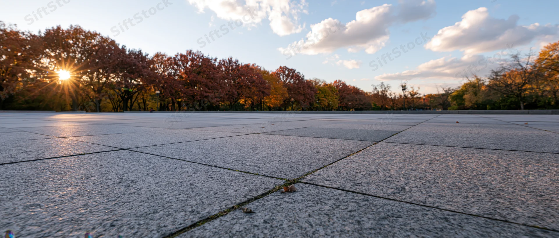 Empty granite-tiled plaza stretching to horizon at sunset with autumn trees in background
