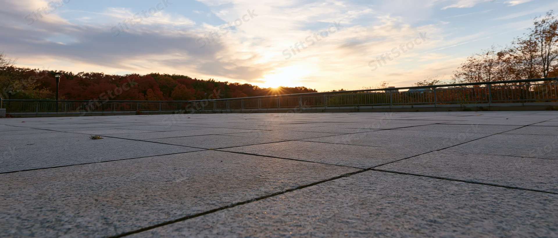 Empty granite-tiled plaza at sunset with autumn trees and metal railing in the distance