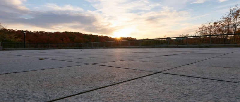 Empty granite-tiled plaza at sunset with autumn trees and metal railing in the distance