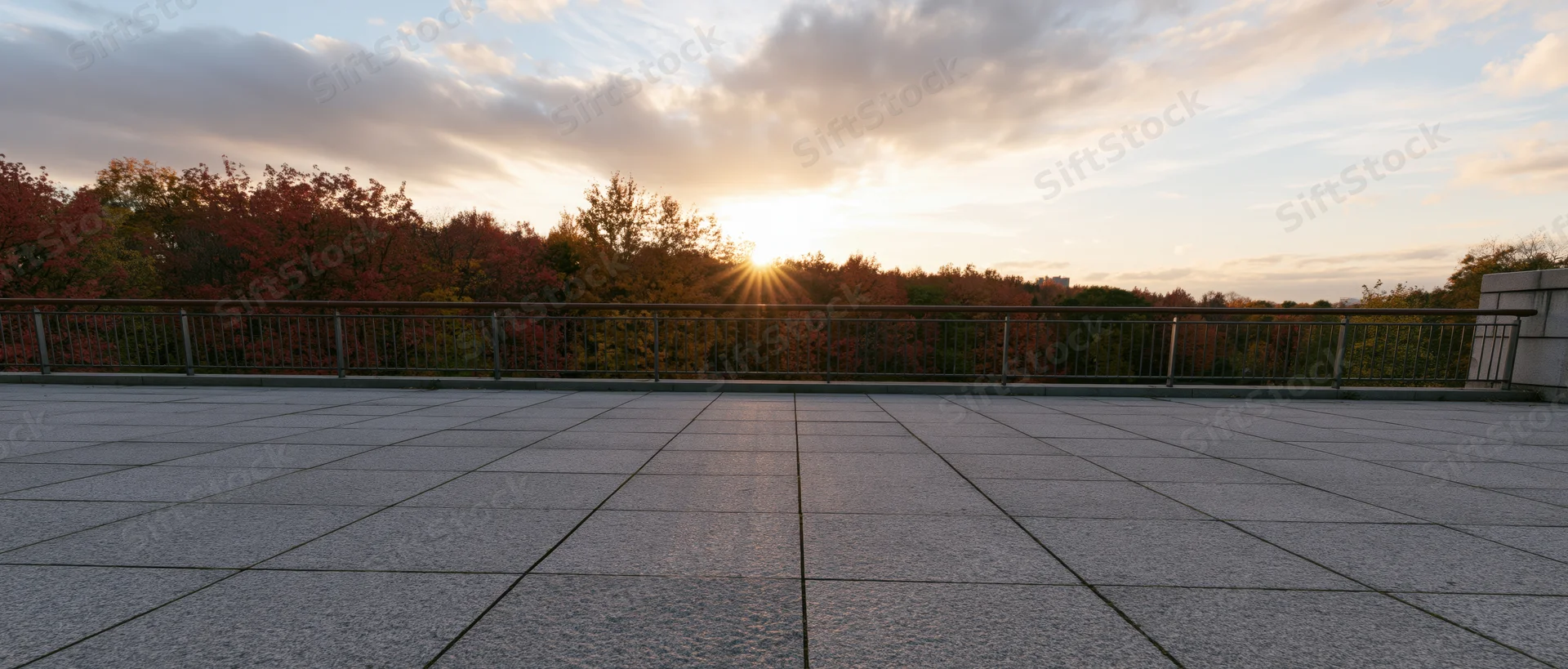 Empty granite-tiled plaza at sunset with autumn trees beyond metal railing
