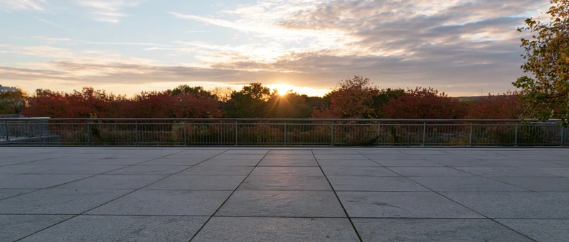 Empty granite-tiled plaza at sunset with autumn trees and metal railing in the distance