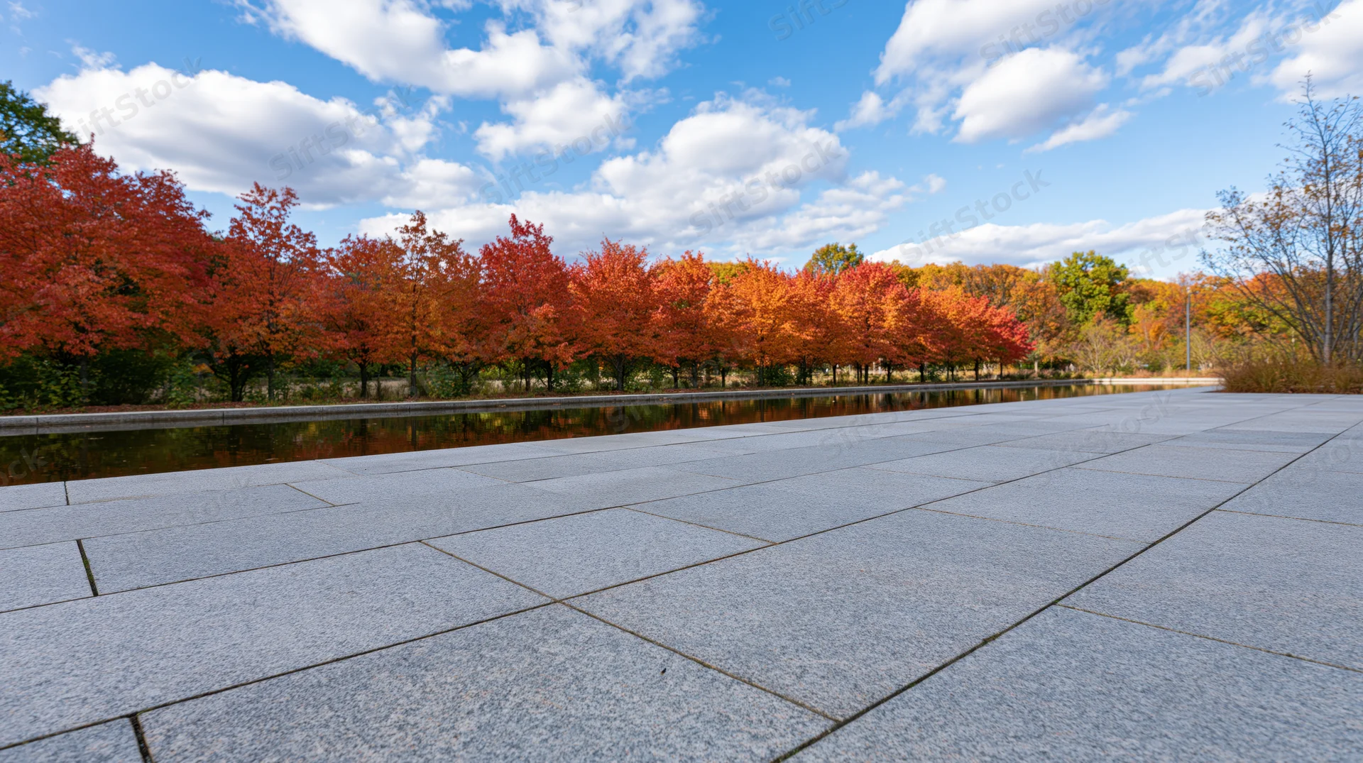 Grey granite plaza stretching toward a line of autumn trees reflected in a calm water feature under a blue sky with clouds