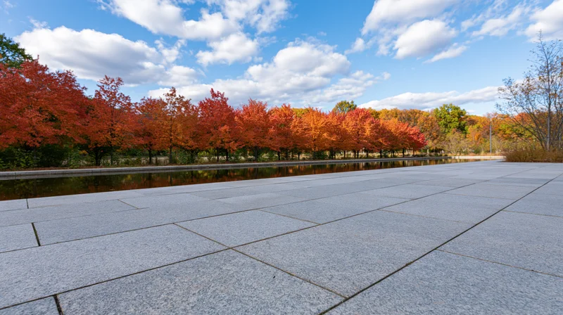 Grey granite plaza stretching toward a line of autumn trees reflected in a calm water feature under a blue sky with clouds