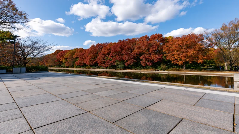 Empty granite plaza leading to autumn trees reflected in a canal under a blue sky
