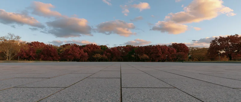 Wide view of a large stone-tiled public plaza stretching toward autumn trees under a cloudy sky at sunset
