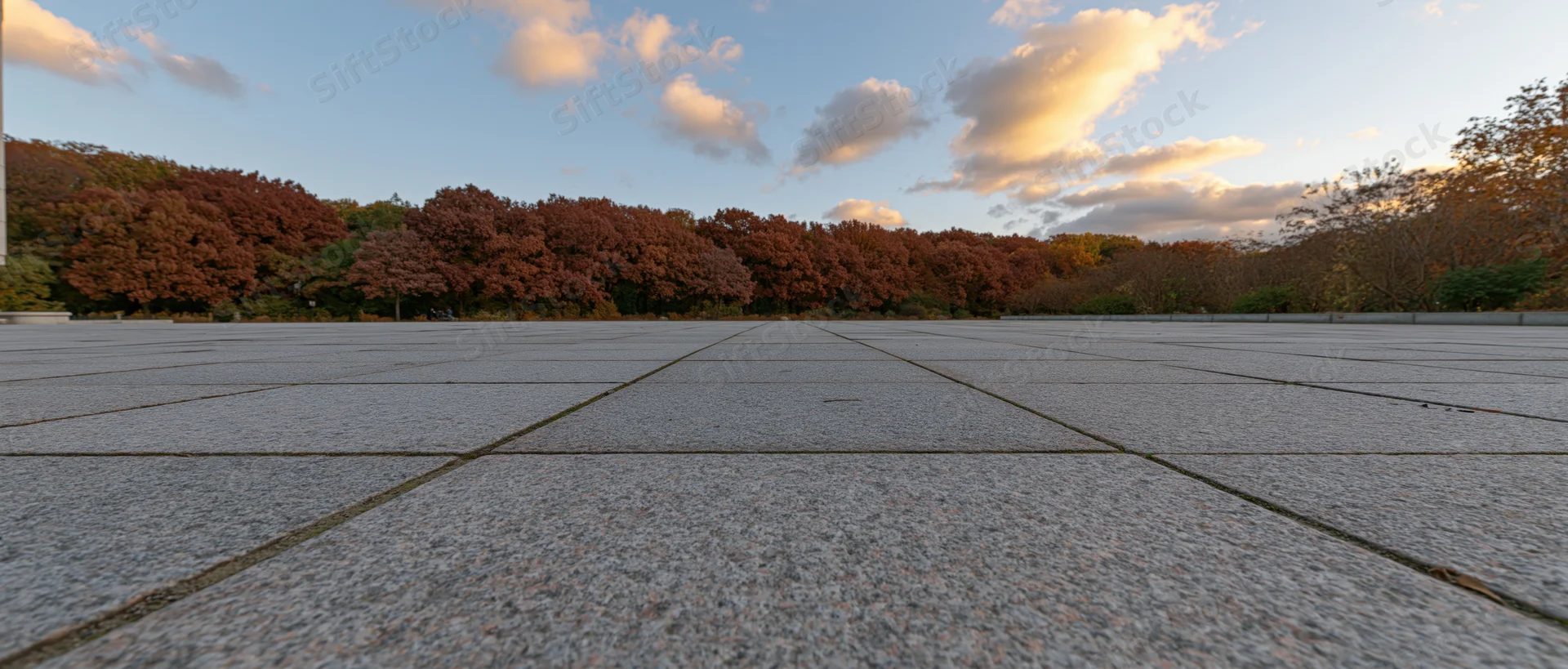 Wide stone-tiled plaza leading to autumn trees under sunset sky