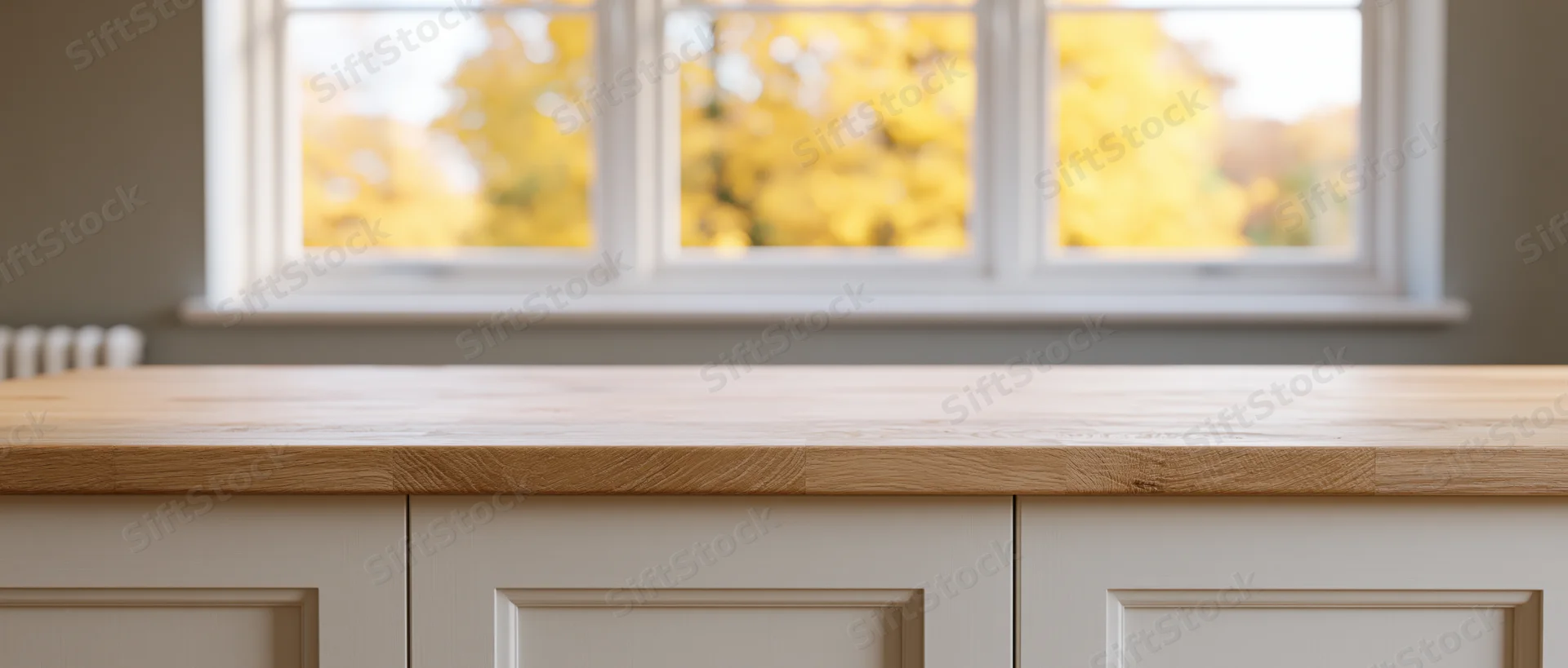 Empty light oak kitchen countertop in front of white cabinets with autumn trees visible through window