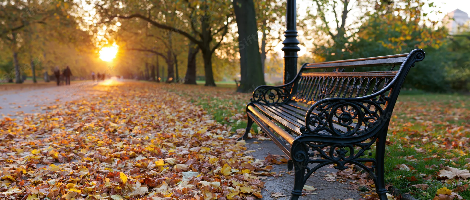 Ornate cast-iron park bench beside a lamppost on a leaf-covered path at sunset