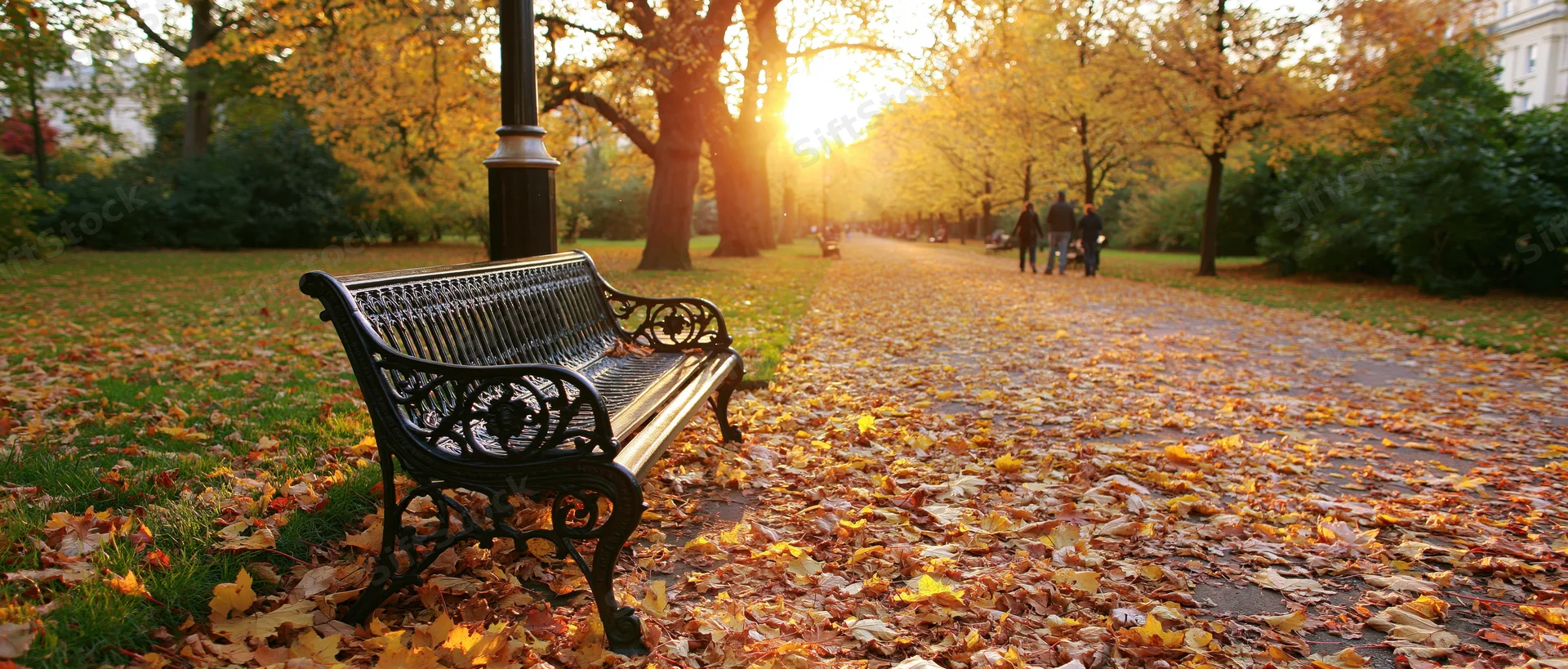 Ornate cast-iron bench beside a lamppost on a leaf-covered park pathway at sunset