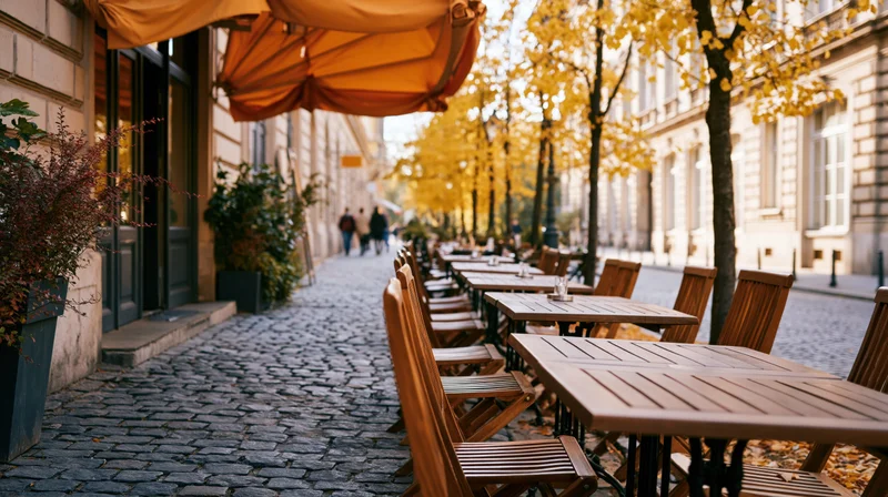 Empty outdoor cafe terrace with wooden tables and chairs along a cobblestone street lined with autumn trees