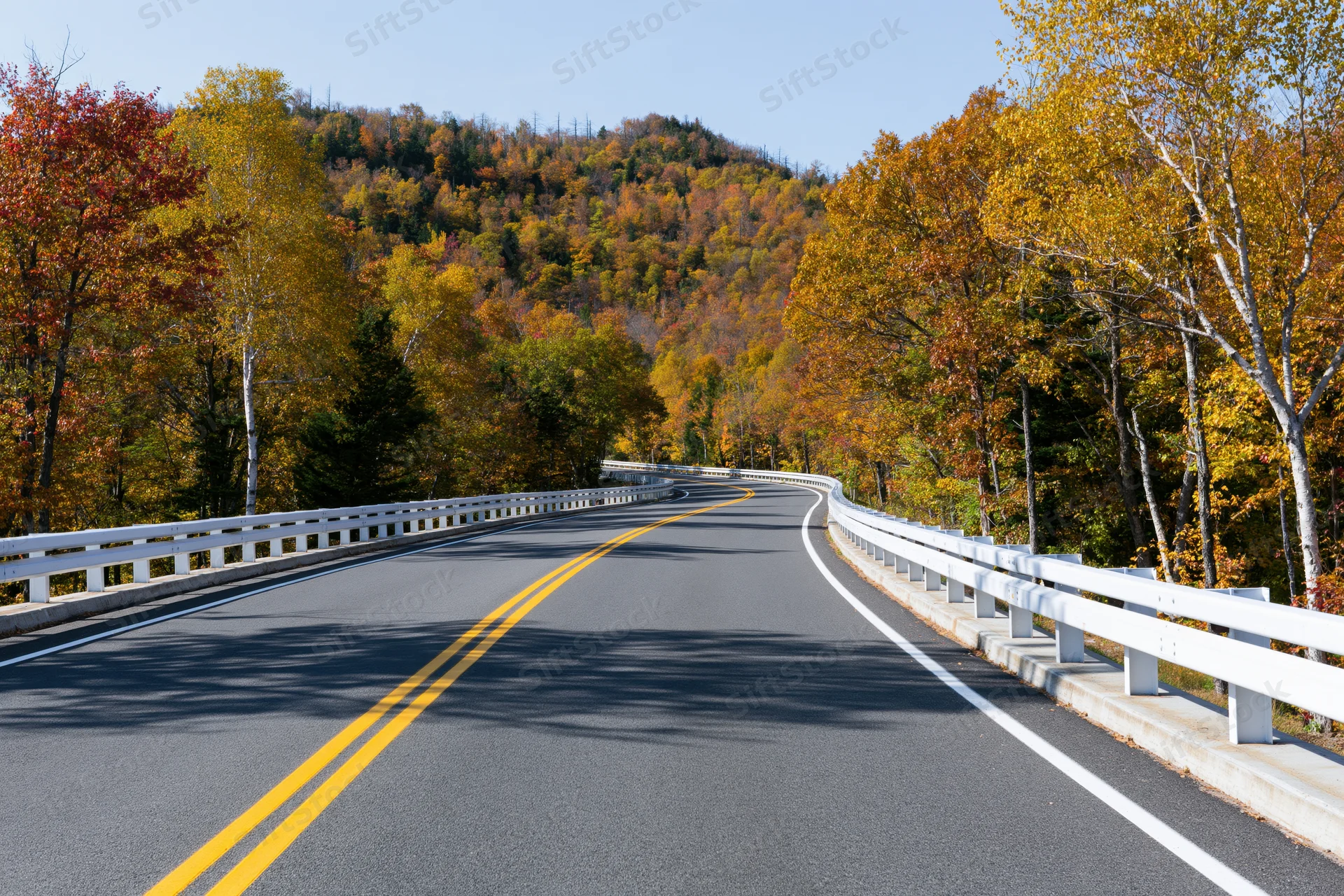 Winding asphalt highway curving through autumn forest with colorful foliage and white guardrails under clear blue sky