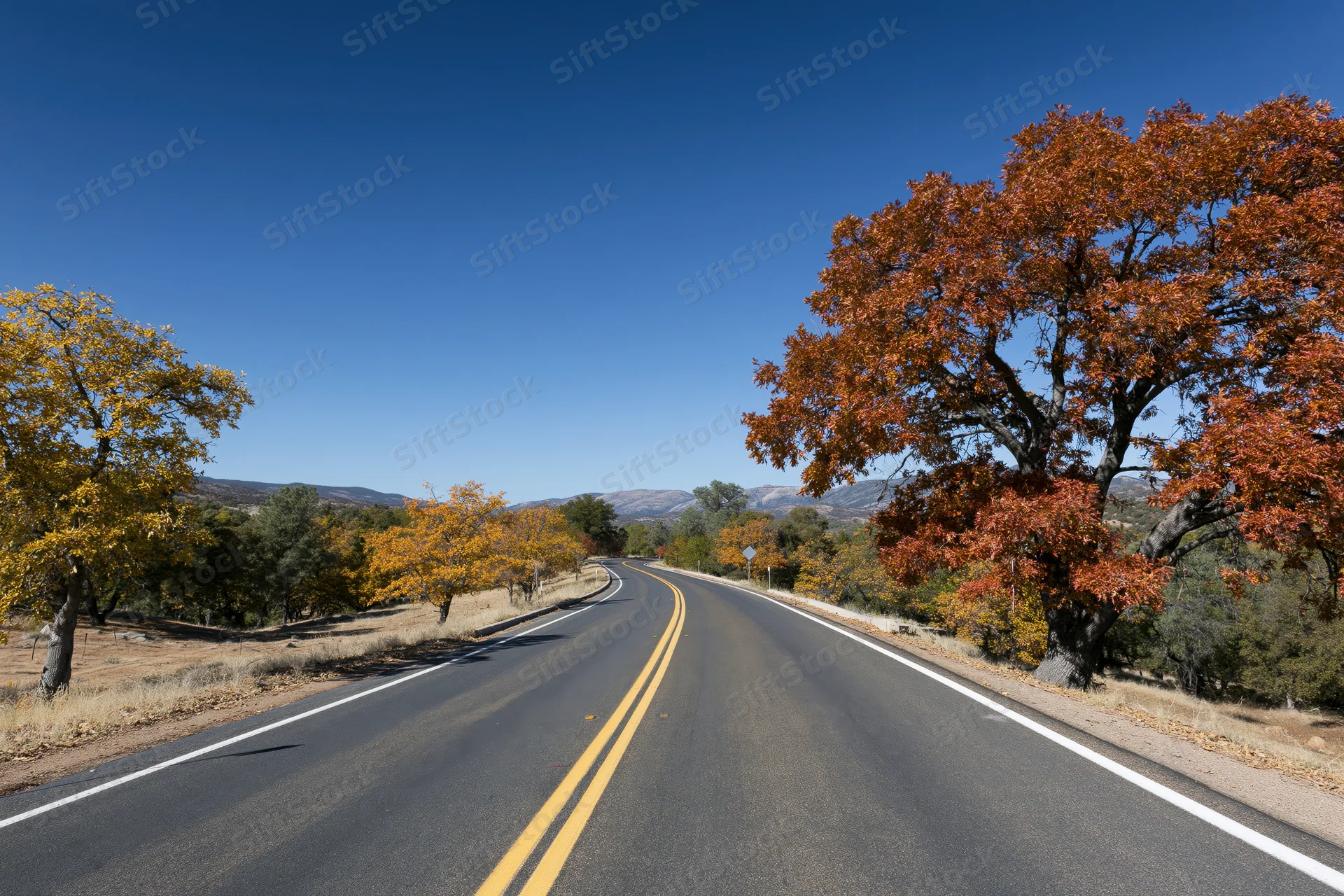 Winding two-lane asphalt road curving through autumn landscape with colorful trees and distant mountains under clear blue sky