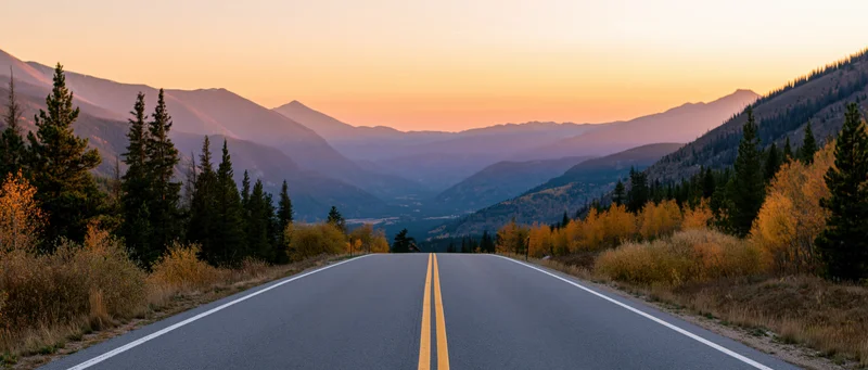 Empty two-lane highway stretching toward distant mountains at sunset through autumn forest