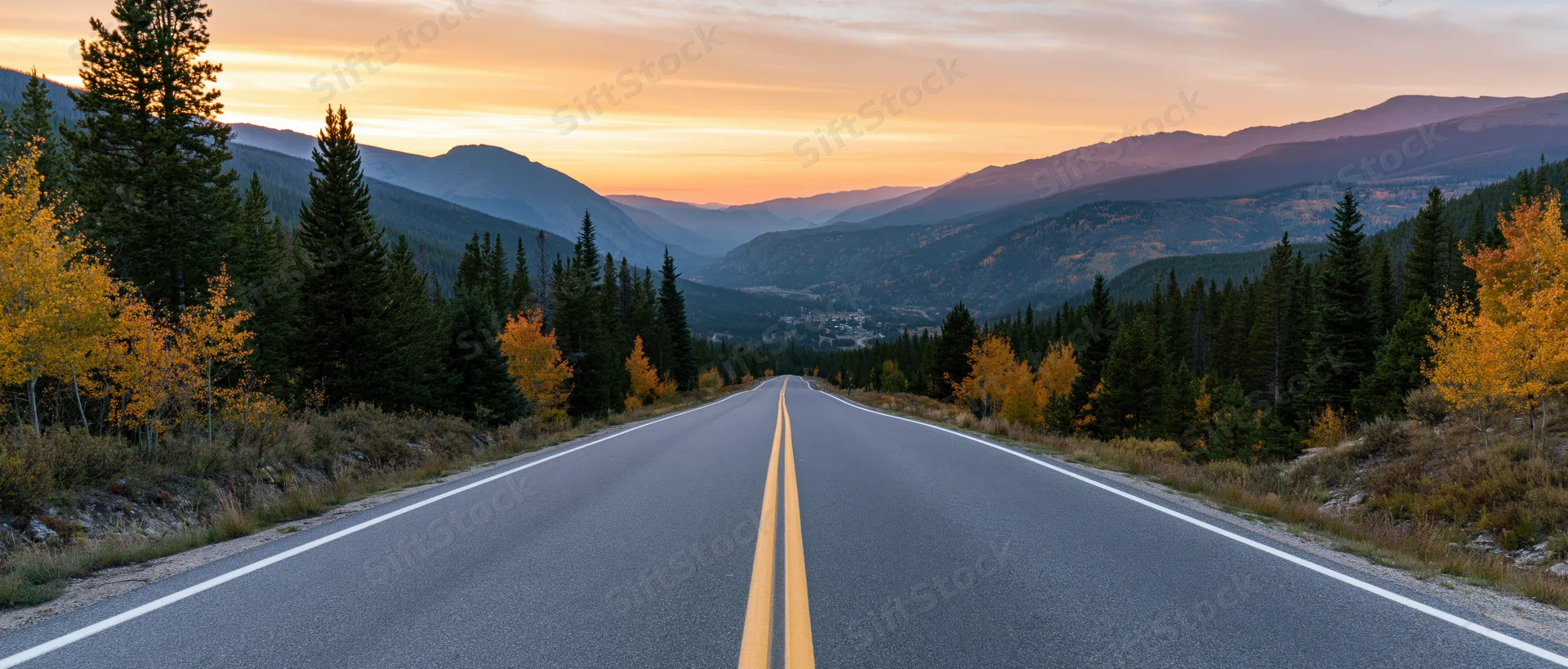 Two-lane highway stretching toward distant mountains at sunset through autumn forest