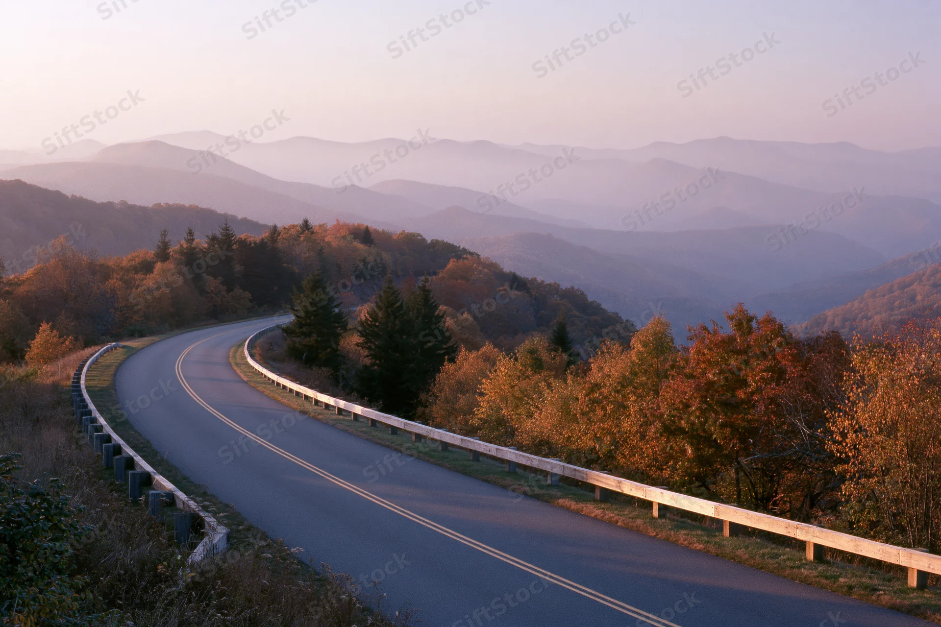 Winding two-lane road curves through autumn forested mountains at sunset