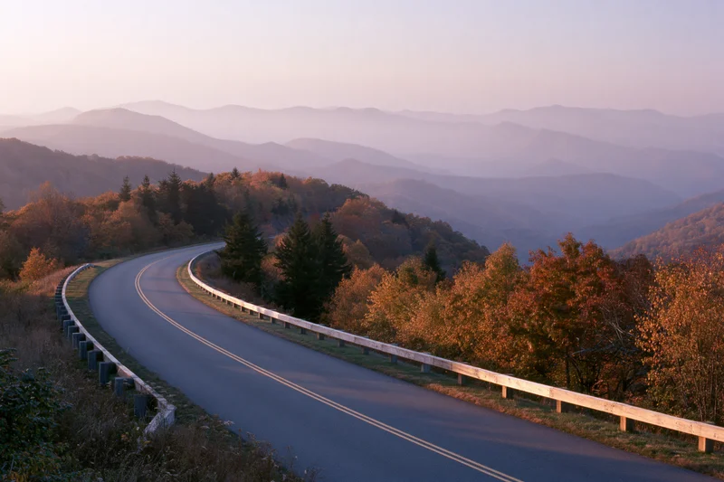 Winding two-lane road curves through autumn forested mountains at sunset