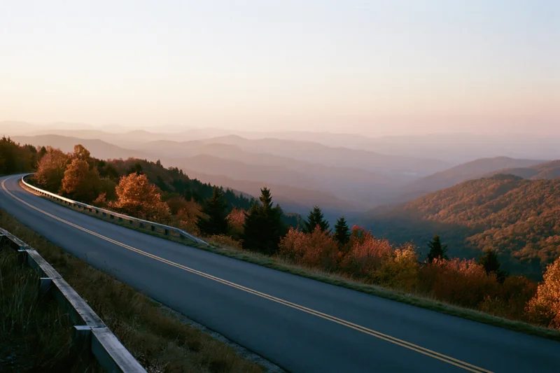 Winding two-lane road curves through autumn forested mountains at sunset