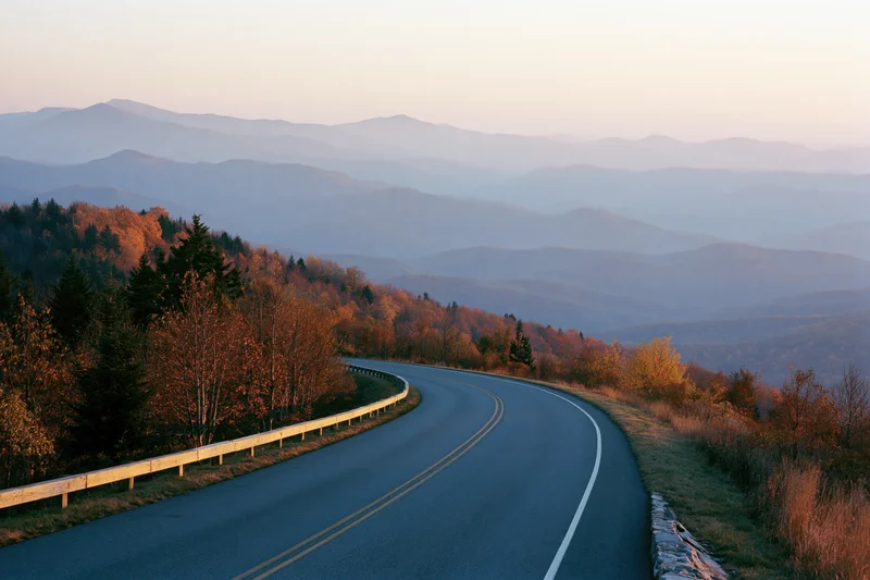 Winding two-lane road curves through autumn forested mountains at sunset