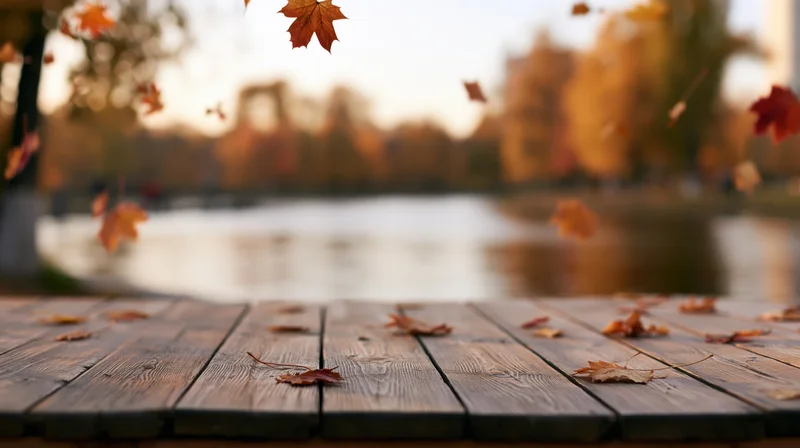 Weathered wooden table surface in foreground with falling autumn leaves and blurred park lake background