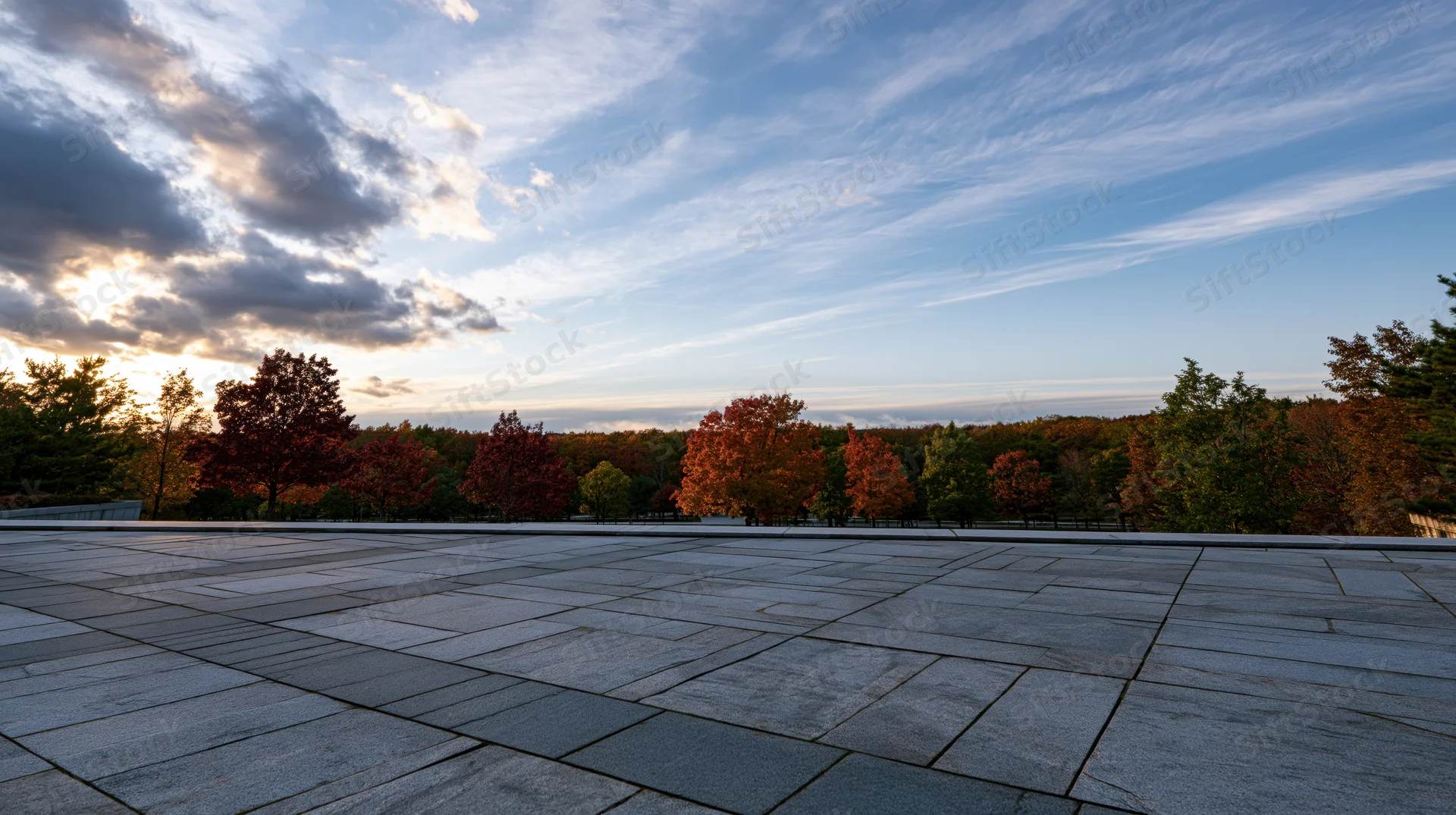 Wide granite tile plaza stretching to horizon with autumn trees and cloudy sky at sunset