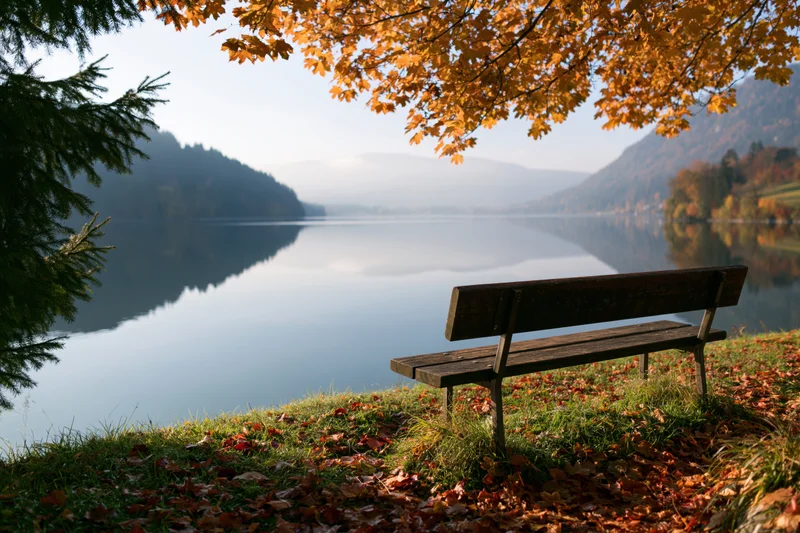 Wooden bench beside calm lake in autumn landscape with falling leaves and mountain reflections