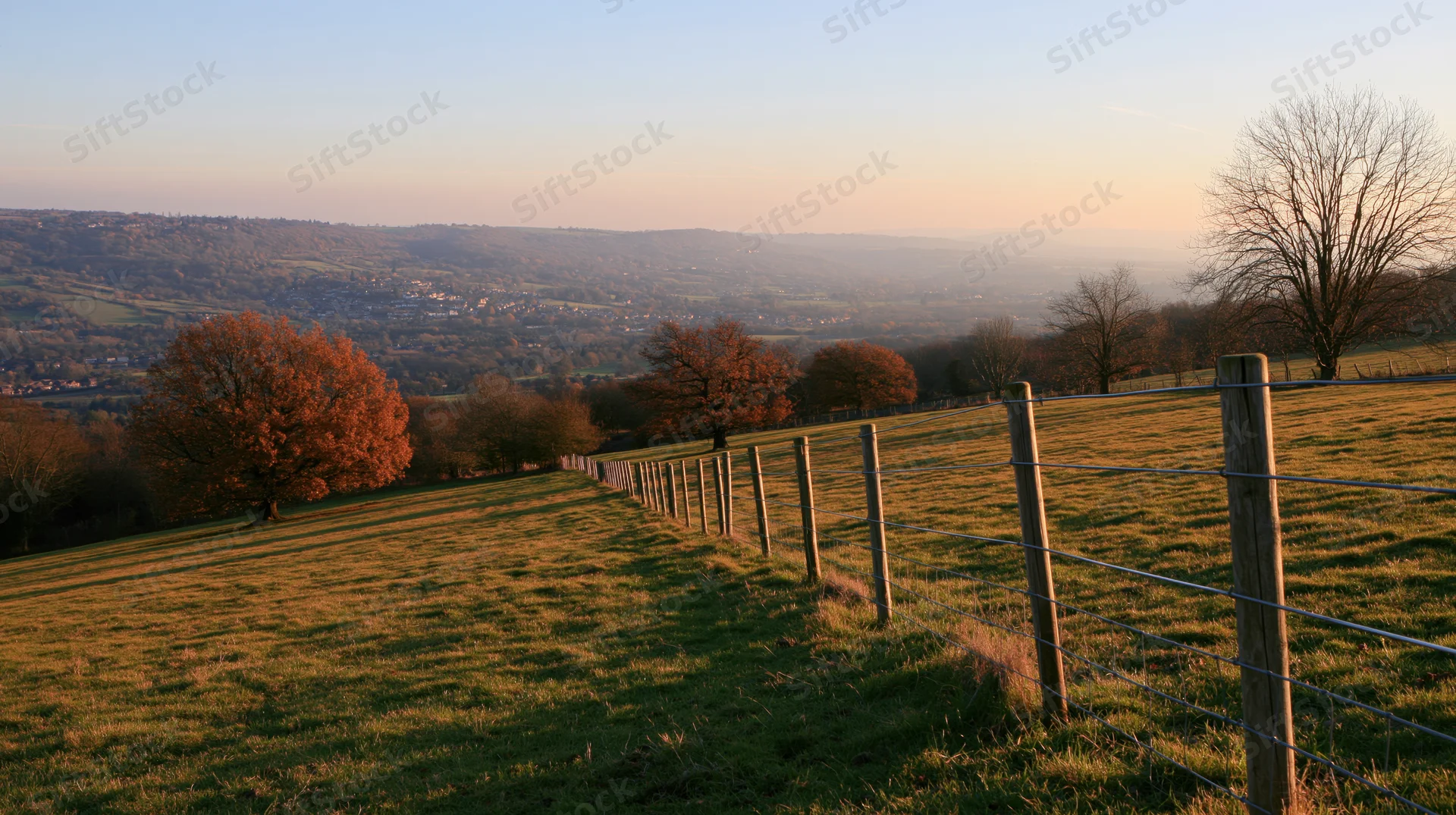 Wide view of a grassy hillside with a wire fence and autumn trees overlooking a valley in the English countryside at sunset